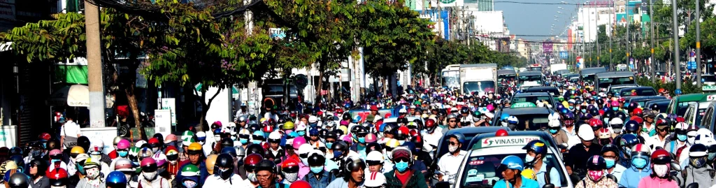 A street is full and crowded with drivers on motor bikes and cars, most wear masks and helmets.