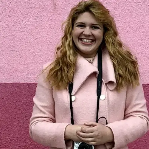 Student in pink jacket in front of pink wall with a camera.