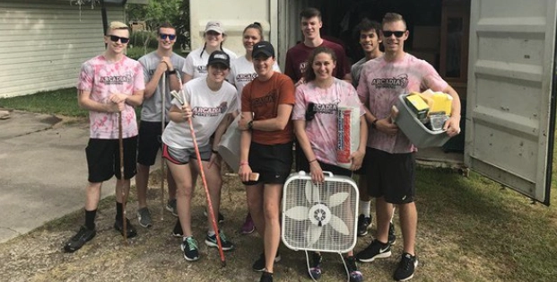 10 students, most of whom wear Arcadia t-shirts, stand together holding a large fan, a box of books, and gardening hoes