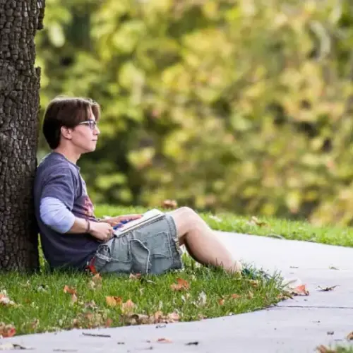 A student sitting against a tree, and doing work while looking around at nature.