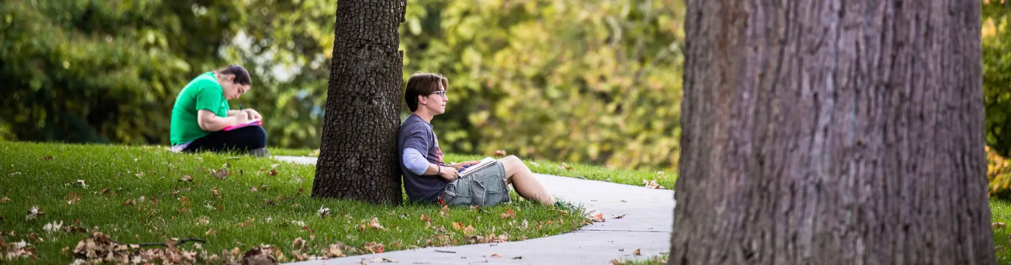 A student sitting against a tree, and doing work while looking around at nature.