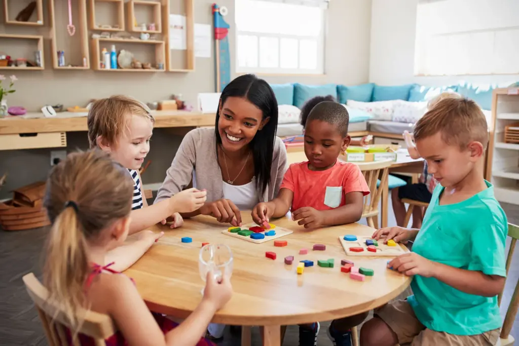 Teacher sitting at table in classroom with four students working on a puzzle with shapes