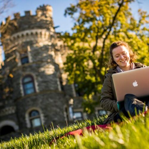A student on her laptop sitting in the grass in front go Grey Towers Castle