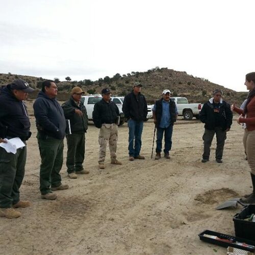 People standing on dirt listening to a speaker in front of them