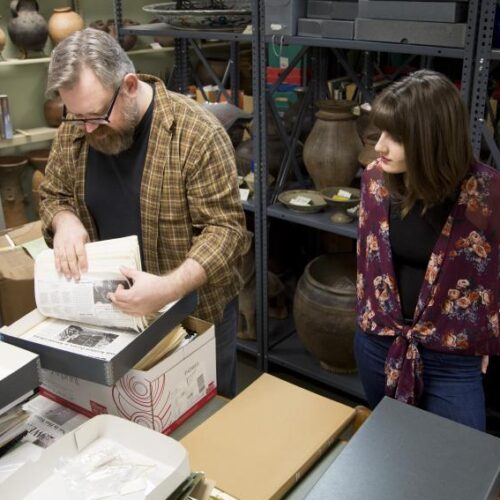 A professor and student look through boxed files in a room with artifacts on crowded shelves
