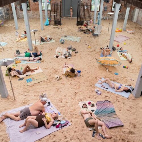 The ground is covered in sand on which people sunbathe on towels