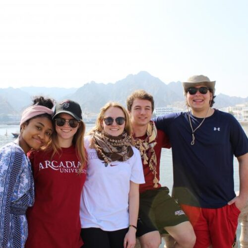 A group of five students with their arms around each other in front of a body of water with mountains in the background