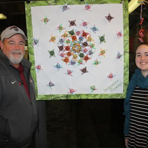 A man and woman stand on either side of a square, geometric art piece with white background and green, leaf pattern border