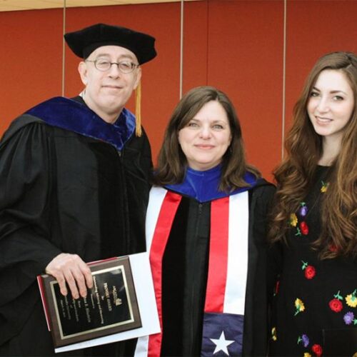 Student (on the right) standing with two people wearing robes and receiving awards