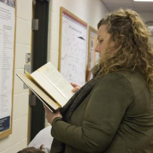 A woman with light curly hair pulled back wears a coat and reads an open book while standing in front of a bulletin board