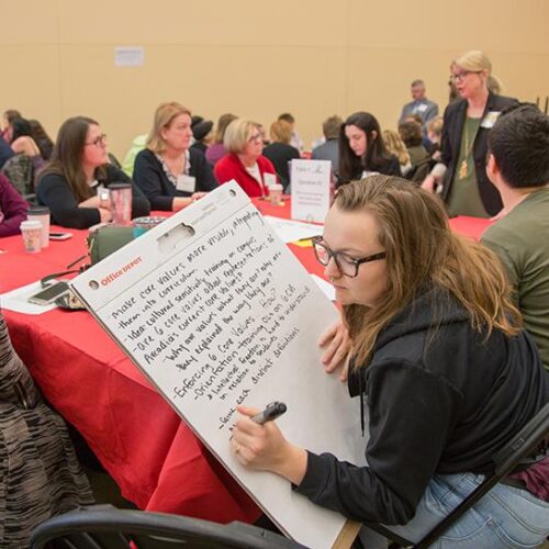 A group of adults sits around a large table with a red tablecloth while one woman in the foreground writes with a marker on a large pad of paper