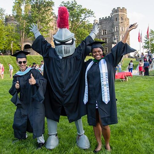 Two students in graduation caps and gowns stands with Arcadia knight mascot