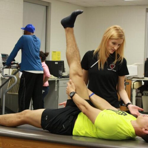 Arcadia Physical Therapy student stretches the leg of a male patient. Another male patient is on a treadmill behind them with two therapists watching a monitor.