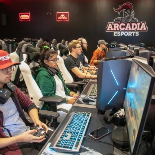 Students are seated in front of monitors with game controllers in their hands, the Arcadia Esports logo on the back wall of the room