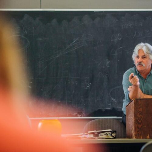 A professor leaning on a podium in front of a chalkboard