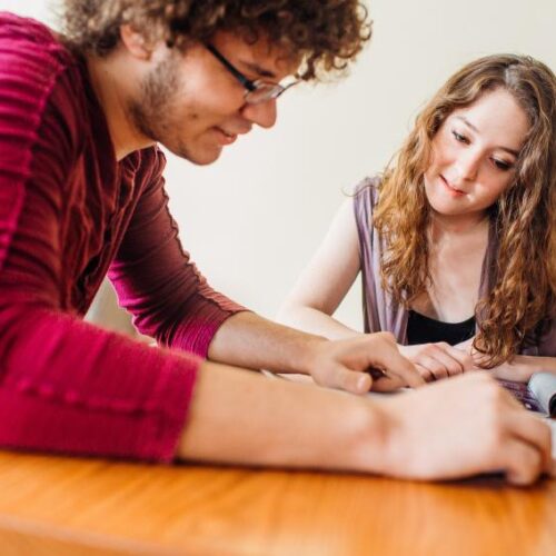Two students look at a notebook and textbook while seated at a table