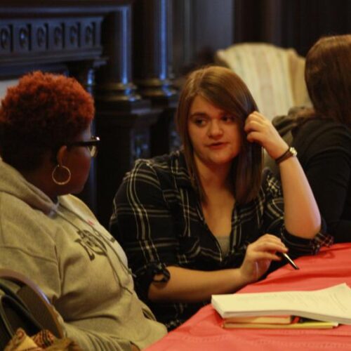 Several people sit at red-covered tables with papers and pen talking to each other