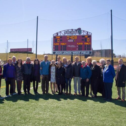 A large group wearing coats stand in front of the scoreboard and sign for Jean Lenox West Field on a sunny day.