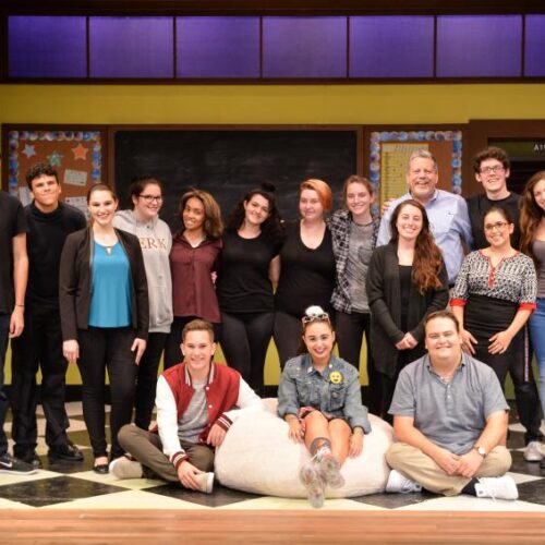 Arcadia Theater students stand in a line with three seated on the checkerboard floor and smile at the camera