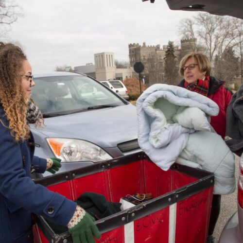 A woman takes a comforter out of the trunk of her car and puts it into a red bin wheeled by another woman in a blue coat