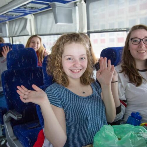 Young female students wave while seated on a bus.