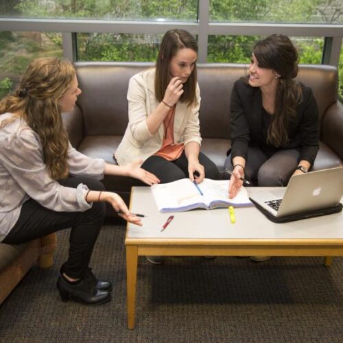Three students sit on a loveseat and chair in the library, a laptop and textbook in front of them