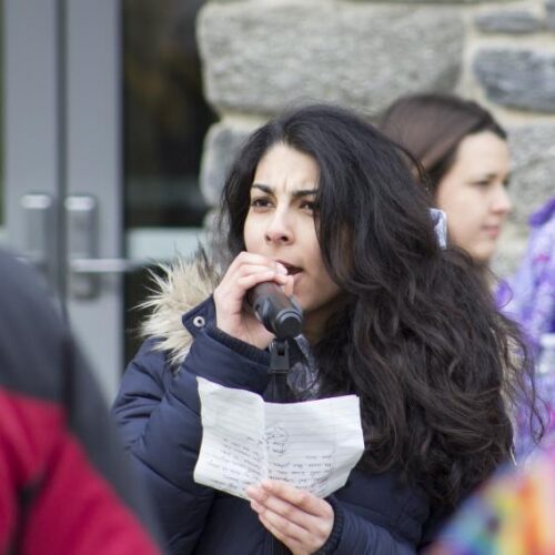 A woman wearing a black parka speaks into a microphone, a piece of notebook paper in her hand