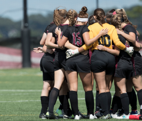 Female athletes wearing tall black socks, cleats, and jerseys, huddle together on the field