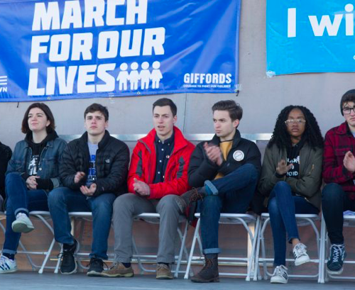 Young people are seated and clapping beneath a large March For Our Lives banner.