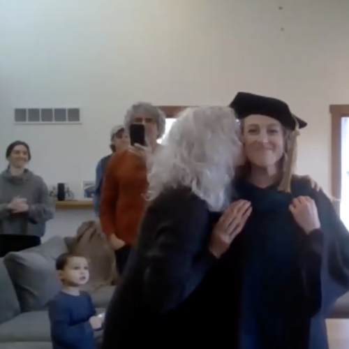 A student in a black cap and gown is kissed on the cheek by a woman with white hair while other people smile at them in the background