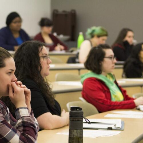 Female students sit attentively at tables in a large classroom