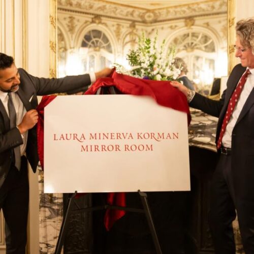 The president of Arcadia University and another man in a suit unveil a sign that says "Laura Minerva Korman Mirror Room" while standing in a room of mirrors