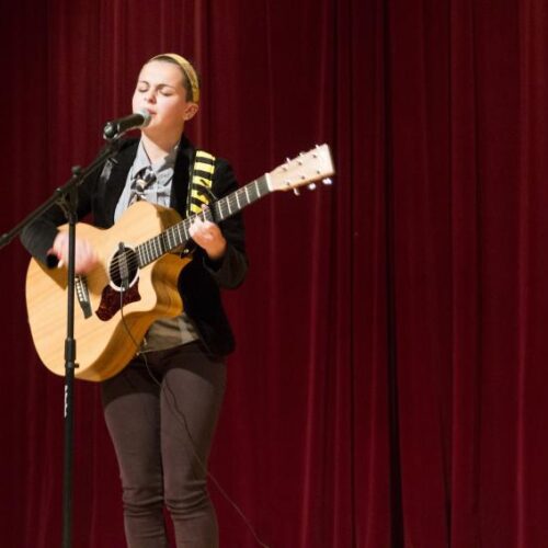 A student stands on stage with red velvet curtains playing guitar and singing into a microphone