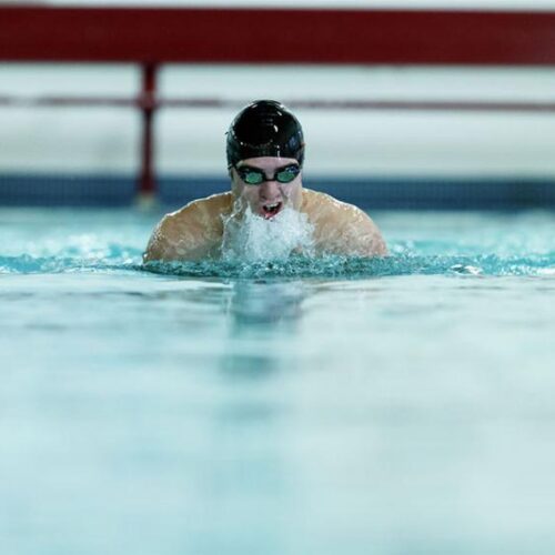 A male swimmer with cap and goggles breaching water's surface during a lap.