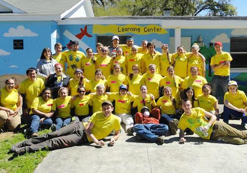 Arcadia Alumni, staff, and students grouped in front of Learning Center Mural in matching yellow shirts.