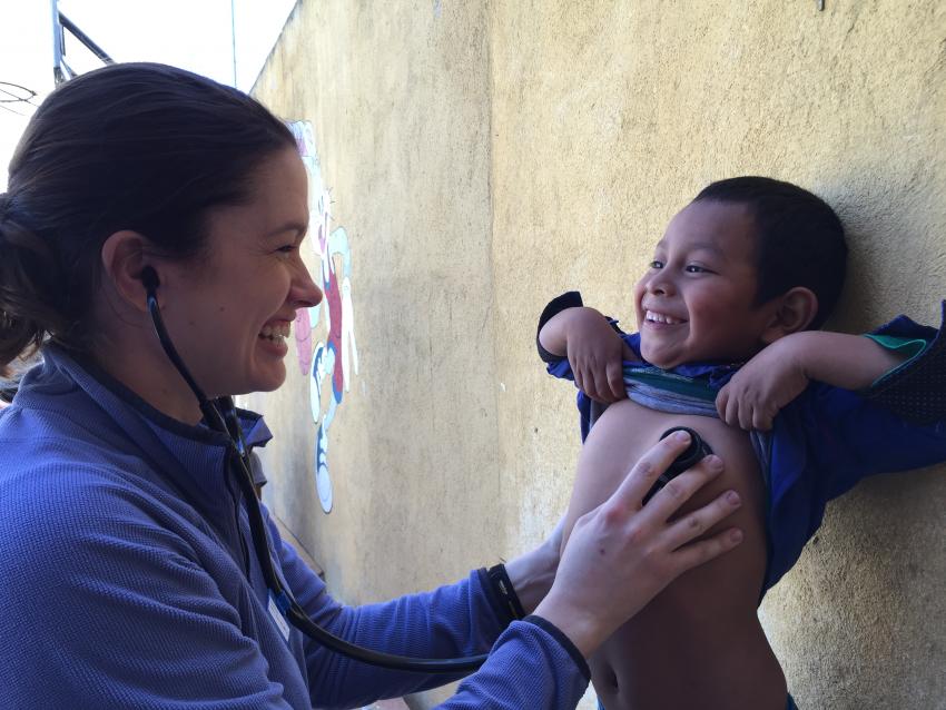 A woman with a stethoscope in her ears presses it against the chest of a small boy lifting his shirt and smiling