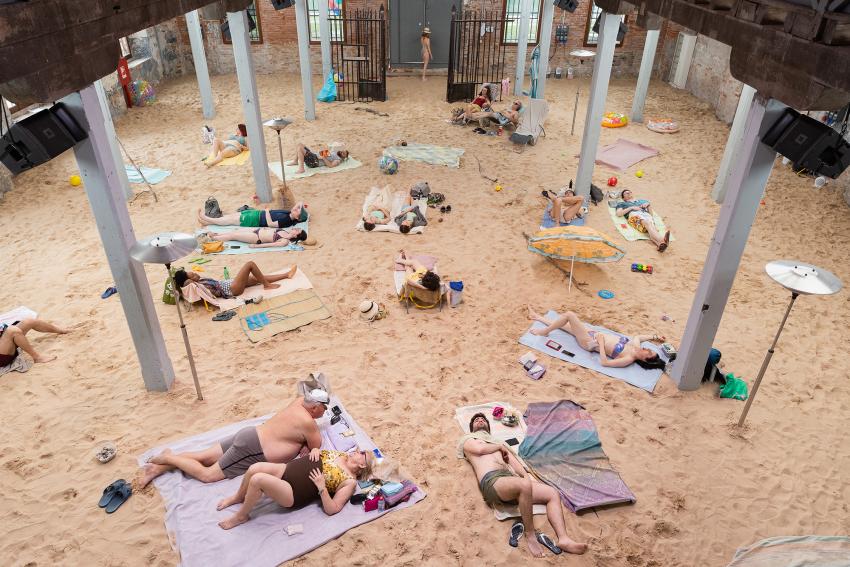 The ground is covered in sand on which people sunbathe on towels