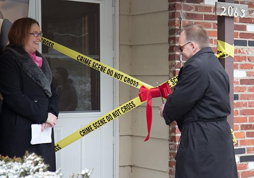 A woman looks on, smiling, as a man cuts a ribbon on two pieces of crime scene tape outside the front door of a house