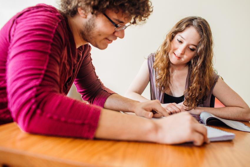 Two students look at a notebook and textbook while seated at a table