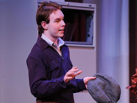 Person speaking on stage with a hat in their hand.