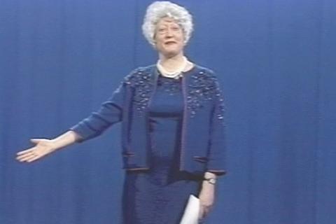 A woman with gray hair, dressed in all blue, looking out at an audience while raising her right hand and holding papers in her left hand.
