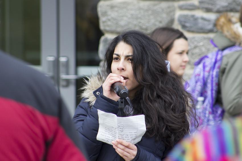 A woman wearing a black parka speaks into a microphone, a piece of notebook paper in her hand