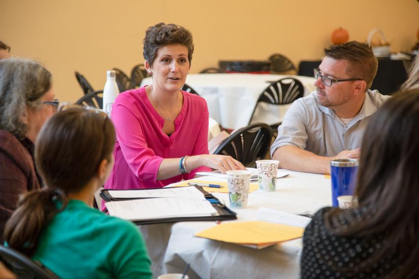 Five people around a table, four looking at a woman in a pink shirt who is speaking