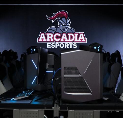 A dark room contains the Arcadia Esports logo on the back wall, with chairs in front of monitors in the foreground