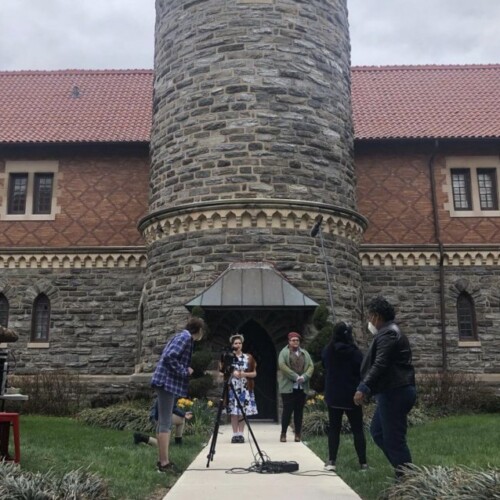 A film crew and cast are shooting outside of a building on campus