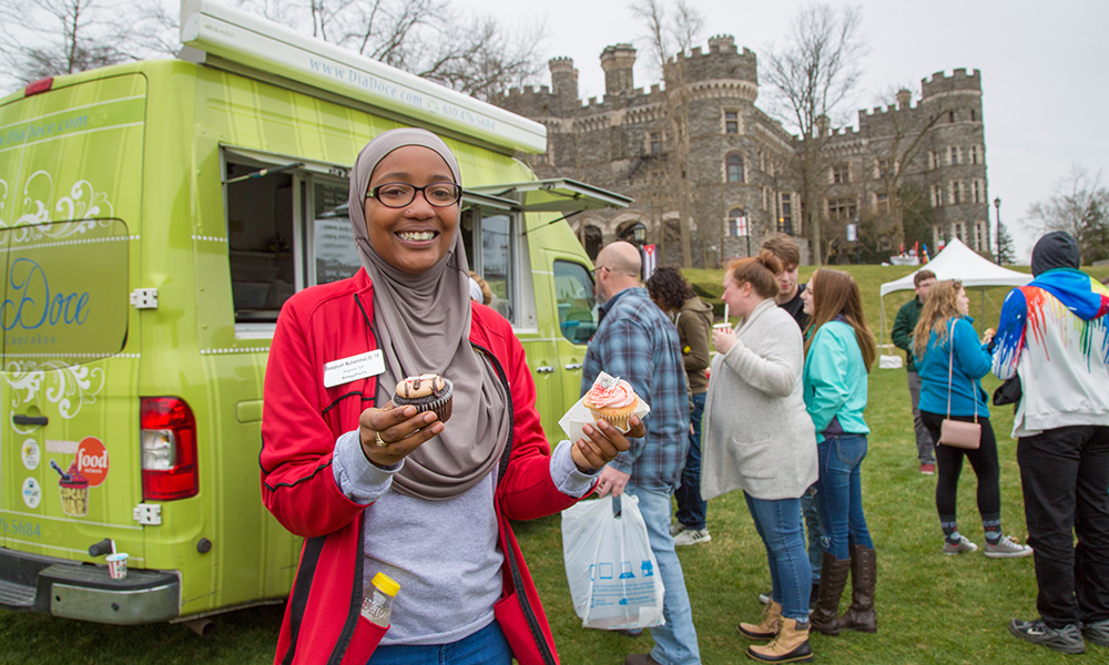 Undergraduate students smiling at the camera while holding two cupcakes in front of the food truck. In the background, people are lined up at the food truck.