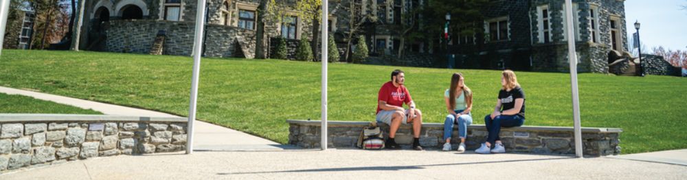 Three students seated outside of Arcadia University on a stone entranceway wall