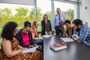 Students and faculty meet around a large table indoors with windows in the background.