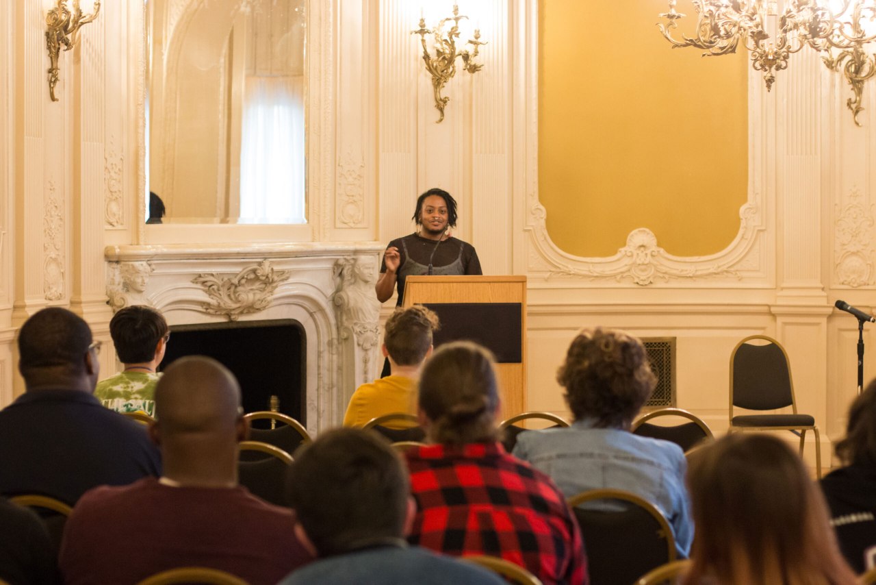 A person at the podium speaking to a group of students in the Mirror Room.