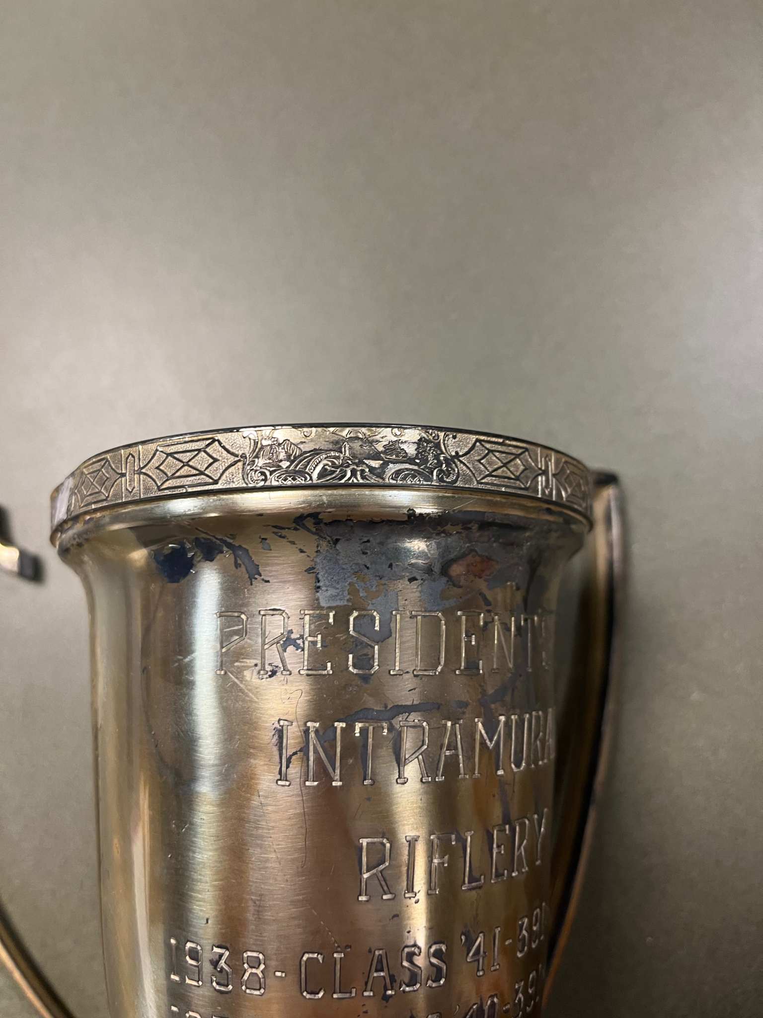 A close up view of an aged silver trophy engraved with "President’s Cup Intramural Riflery Trophy dating from 1938-1948."